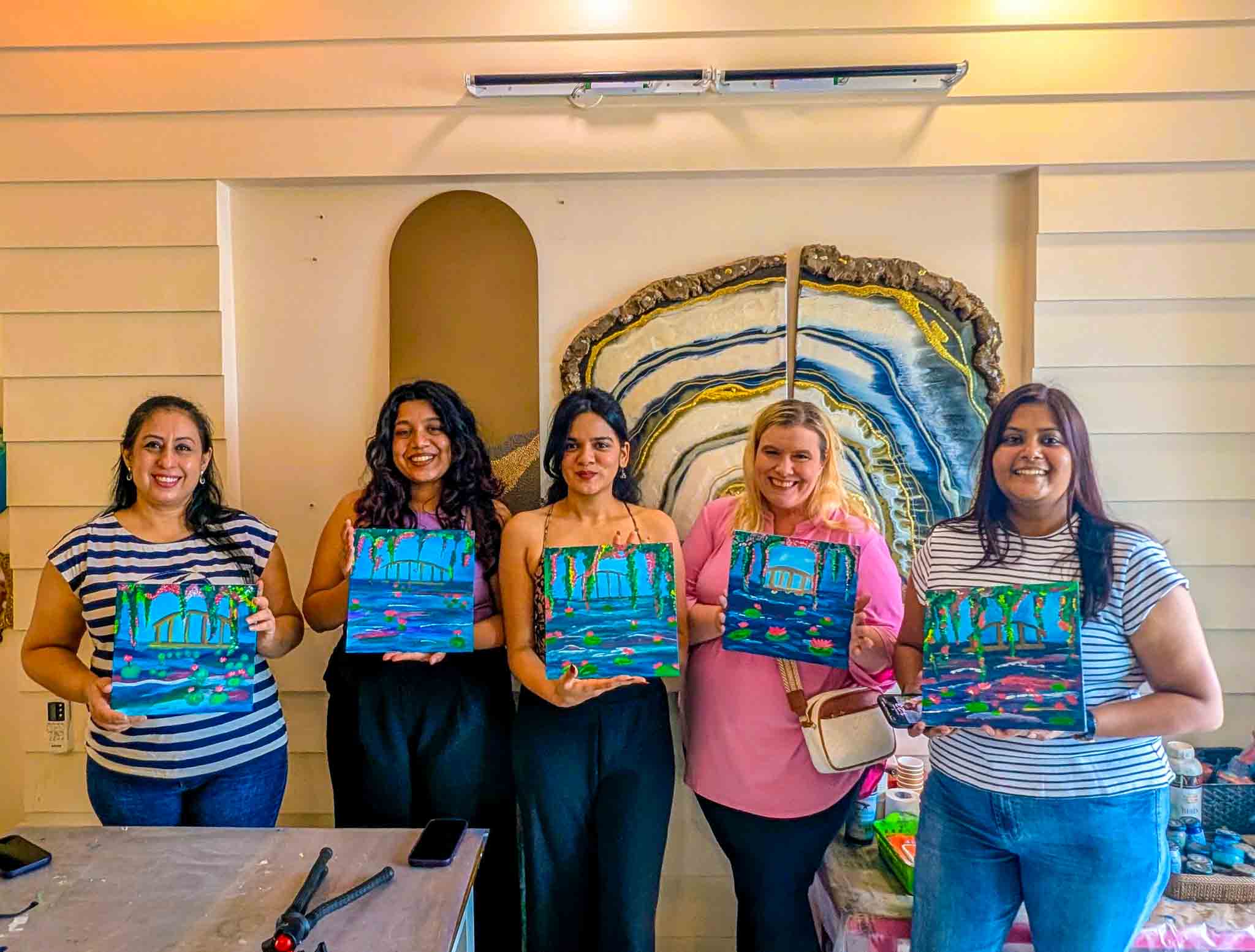 Five women proudly hold their beginner-friendly painted canvases featuring a bridge and water lilies during a Sip & Paint session.